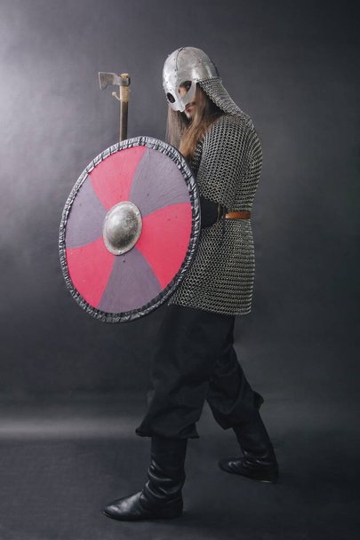 Viking staat met een bijl op een grijze achtergrond. Een man met een maliënkolder en een helm heeft een wapen. Middeleeuwse krijger in harnas. Studiofoto - Foto, afbeelding