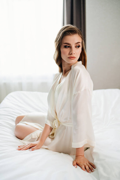 beautiful woman with makeup, long hair sits in white robe on bed - Photo, Image