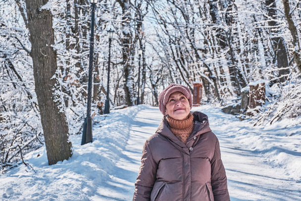 Kahverengi ceketli, pembe kürklü yaşlı bir kadının portresi. Kış karlı bir parkın arka planına bakıyor. - Fotoğraf, Görsel