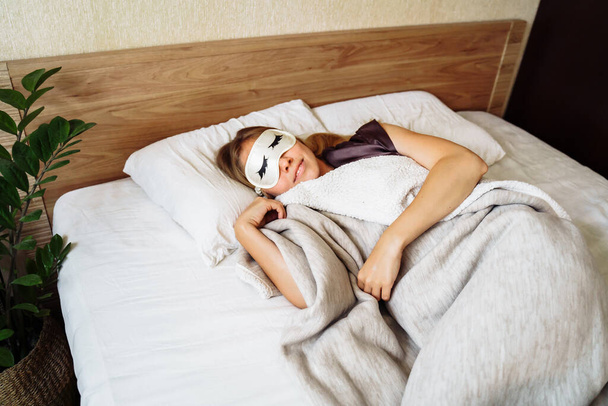 Pretty girl in night robe sleepping sweetly in snow-white bed in her room. View from the top, above. Wearing eyelash-patterned night goggles. Cozy covered with a blanket. Sweet morning dream at dawn. - Photo, Image