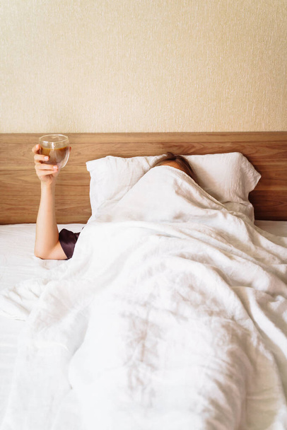 Uykulu genç bir kadın kar beyazı bir yatakta yatıyor. Başında bej bir battaniye vardı. Bir fincan kahveyle elini kaldırdı. Sabah uyanmanın zorluğuyla ilgili komik bir kare.. - Fotoğraf, Görsel