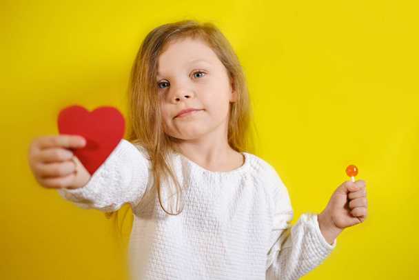 Portrait of smiling kid girl holding a red heart for you and candy in the second hand, isolated on yellow background with copy space. V2 - Fotó, kép