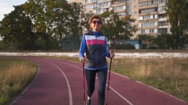Spor sahasında yürüme direğiyle çalışan yaşlı bir kadın. Yaşlılar sağlıklı bir yaşam tarzı. Kıdemli İskandinav yürüyüşü. Şehir stadyumunda koşan bastonlu olgun bir kadın. - Video, Çekim