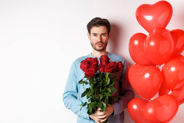 Walentynkowy romans. Pewny siebie młody mężczyzna trzymający bukiet czerwonych róż, stojący obok balonów serc, idący na romantyczną randkę z kochankiem, białe tło - Zdjęcie, obraz