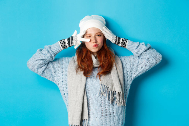 Winter and holidays concept. Happy redhead girl in beanie hat, scarf and gloves showing peace sign and pucker lips sassy, staring at camera, wearing sweater, posing over blue background - Foto, Imagen