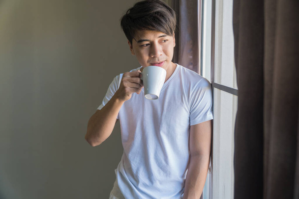 The handsome man stands at the window holding a white cup of coffee in his hand. - Foto, Bild