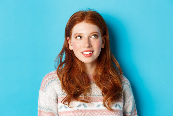 Close-up of dreamy redhead girl imaging something, staring at upper right corner and smiling, standing in winter sweater against blue background - Photo, Image