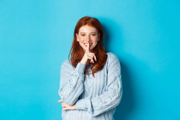 Pretty redhead teenager hushing and smiling, telling a secret, standing in sweater against blue background - Photo, Image