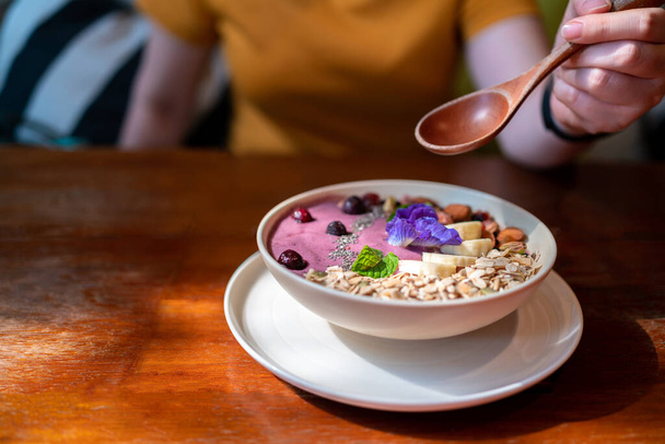 Hand holding wooden spoon over acai bowl mix with blueberry smoothie, fresh banana, berries, sunflower seeds, chia seeds with mint and butterfly pea. Vegan breakfast for healthy and veganism people. - Photo, Image