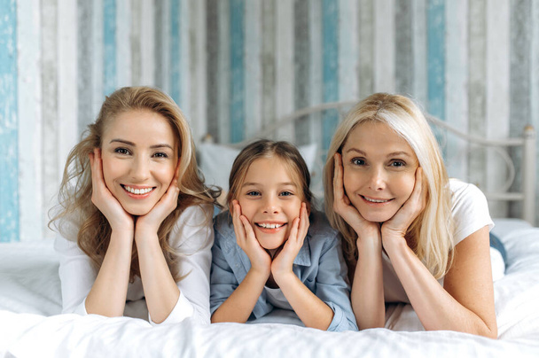 Ritratto di tre generazioni femminili. Felice nonna caucasica, figlia e nipote sono sdraiati a casa sul divano, guardando la fotocamera e sorridendo. Relazioni e valori familiari - Foto, immagini