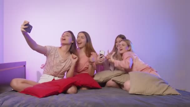 Neljä onnellista nuorta naista yöpuvussa istuu sängyllä, laulaa ja nauhoittaa videota puhelimellaan polttareissa. - Materiaali, video