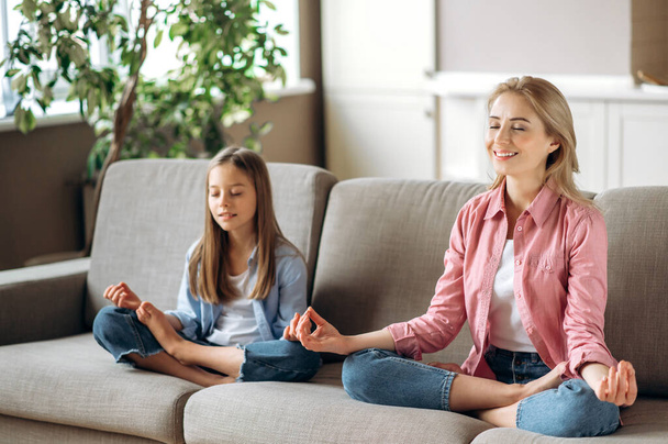 Mom and daughter relationship. Happy caucasian mom and cute little daughter doing yoga and meditation sitting in lotus position at living room on the couch, calmness, relaxing - Foto, Imagen