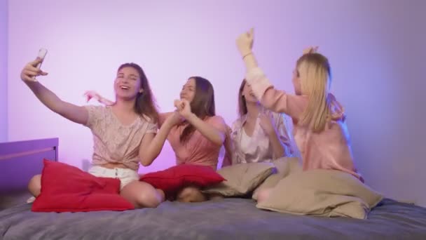 Neljä onnellista nuorta naista yöpuvussa istuu sängyllä, laulaa ja nauhoittaa videota puhelimellaan polttareissa. - Materiaali, video