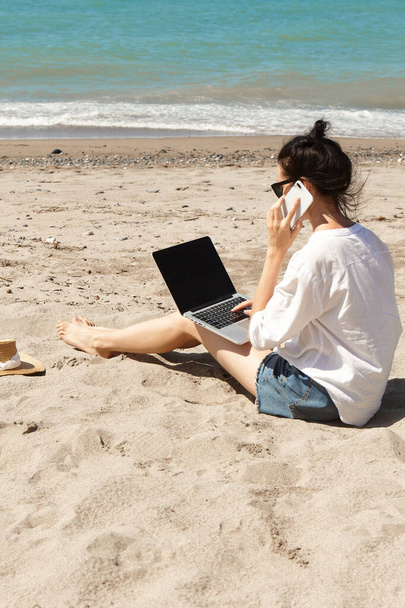 Giovane donna che usa il computer portatile e parla al telefono su una spiaggia. Vacanza stile di vita di comunicazione. Concetto di lavoro indipendente - Foto, immagini