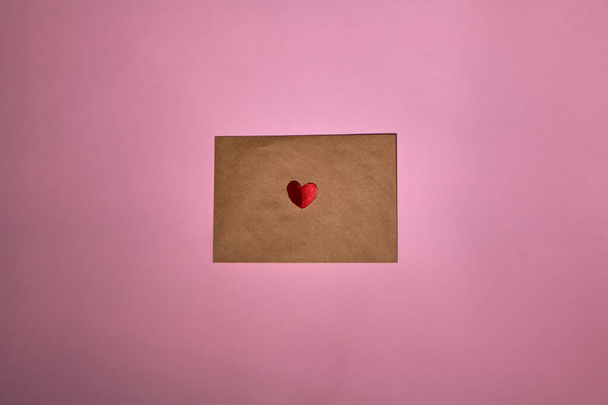 Un cuore rosso giace su una busta marrone per San Valentino. Sfondo rosa con busta e cuore rosso. Lettera d'amore o concetto di messaggio. Piatto, vista dall'alto. Copia spazio per il testo.  - Foto, immagini
