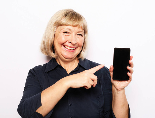 Portrait of a senior woman showing or presenting mobile phone application and pointing finger to smartphone on hand isolated over white background. - Foto, Imagen