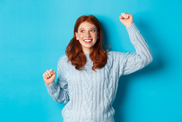 Glückliches rothaariges Mädchen, das für die Mannschaft wurzelt, mit erhobener Hand jubelt und lächelt, Sieg oder Erfolg feiert, vor blauem Hintergrund steht - Foto, Bild