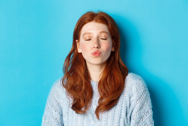 Close-up of young woman with red wavy hair standing with eyes closed and puckered lips, waiting for kiss against blue background - Photo, Image