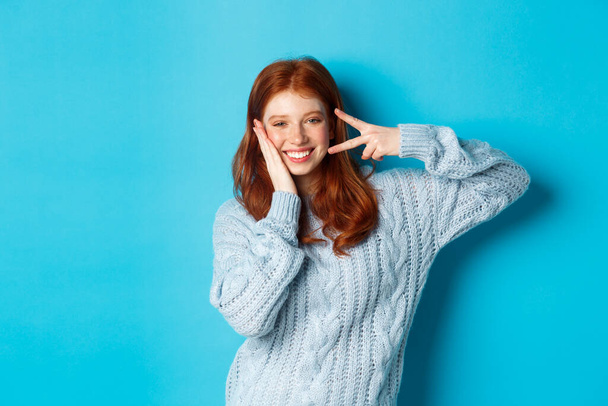 Fröhliche rothaarige weibliche Modell sendet gute Stimmung, lächelt und zeigt Friedenszeichen, steht über blauem Hintergrund - Foto, Bild