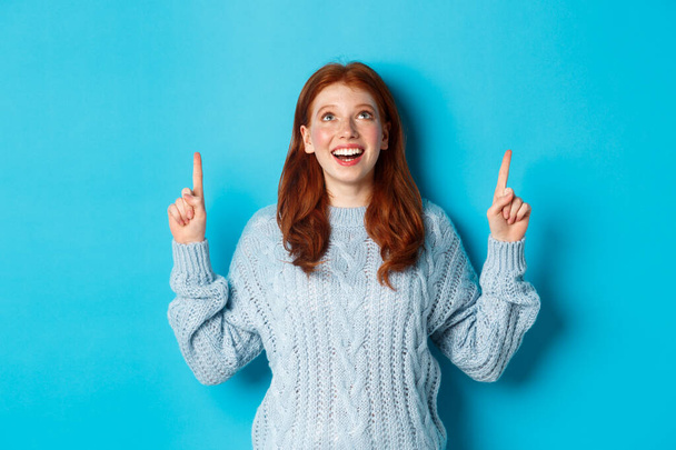 Winter holidays and people concept. Beautiful teenage girl with red hair, wearing sweater, pointing fingers up at logo and smiling, showing advertisement, blue background - Photo, Image