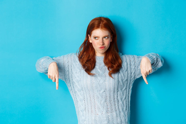 Winter holidays and people concept. Indecisive redhead girl in sweater pointing fingers down and thinking, having doubts, standing over blue background - Foto, immagini