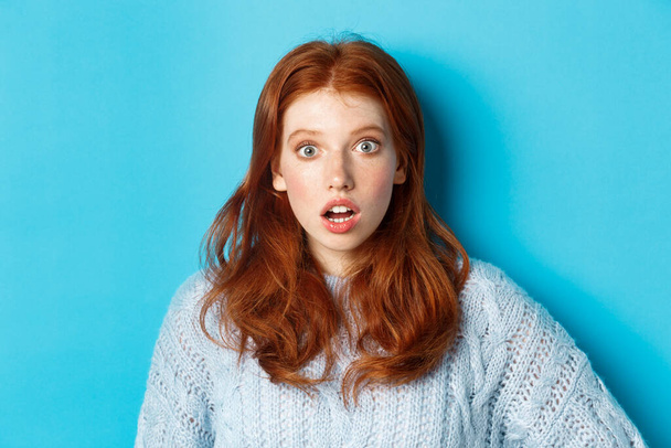 Close-up of startled redhead girl stare at camera speechless, open mouth in awe, standing over blue background - Фото, изображение