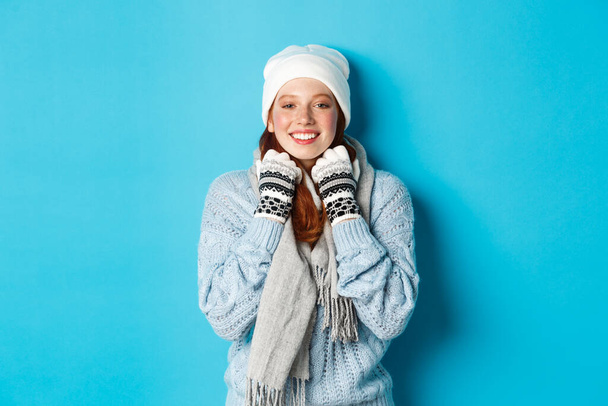 Winter- und Ferienkonzept. Nettes rothaariges Mädchen in weißer Mütze und Handschuhen, das in die Kamera lächelt, erfreut schaut, vor blauem Hintergrund steht - Foto, Bild