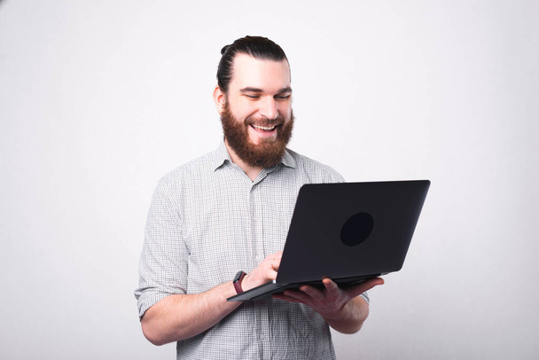 Portrait d'homme barbu heureux en chemise à l'aide d'un ordinateur portable sur un mur blanc - Photo, image