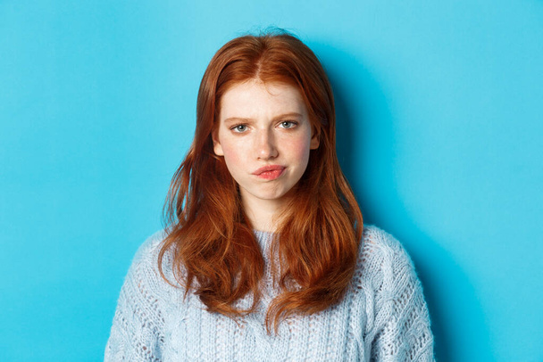 Adolescente déçue aux cheveux roux, fronçant les sourcils et souriant mécontente, regardant vers le jugement, debout sur fond bleu - Photo, image