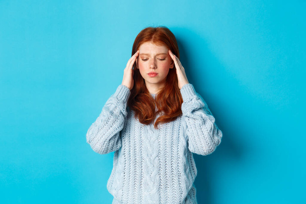 Distressed teenage redhead girl touching head, looking down with troubled face expression, standing against blue background, have problem - Photo, Image