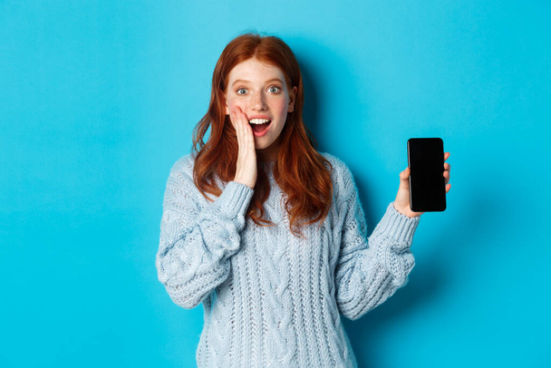 カメラを見て、スマートフォンの画面を見て、モバイルアプリケーションを実証し、青い背景の上に立っている素晴らしい赤毛の女の子 - 写真・画像