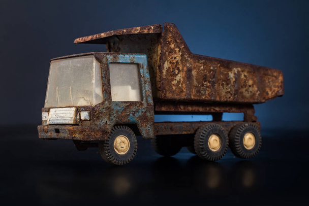 Стара, брудна і дуже іржава модель олов'яного металу вантажівки в синьо-сірому кольорі в чорно-синьому задньому плані на відбиваючій поверхні. Не вистачає одного колеса
. - Фото, зображення