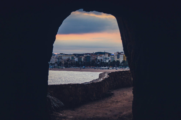 Looking through a rock tunnel on a pedestrian walkway towards sandy beachfront on popular tourist destination Lloret De Mar in Spain in evening with sun slowly falling behind the buildings. - 写真・画像