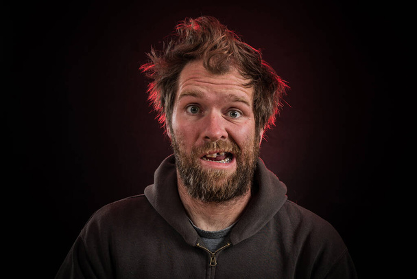 Un uomo goofy con la barba, bretelle dentali su uno sfondo rosso scuro illuminato con cappuccio marrone o maglione e capelli elettrificati. - Foto, immagini