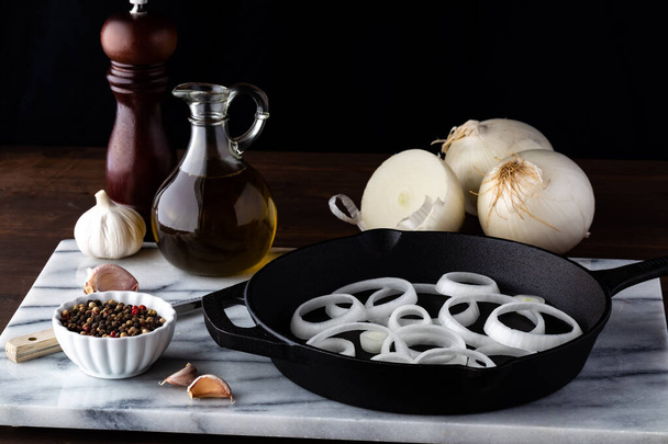 Τηγάνι τηγανιτό χυτοσίδηρο γεμάτο με φέτες λευκού κρεμμυδιού περιτριγυρισμένο από μαγειρικά υλικά. - Φωτογραφία, εικόνα