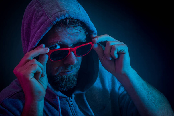 mladý muž pózuje s brýlemi a mikinou přes hlavu v temném dramatickém prostředí s červeným a modrým světlem ze strany. Gang styl policie vyhledávání fotografie. - Fotografie, Obrázek