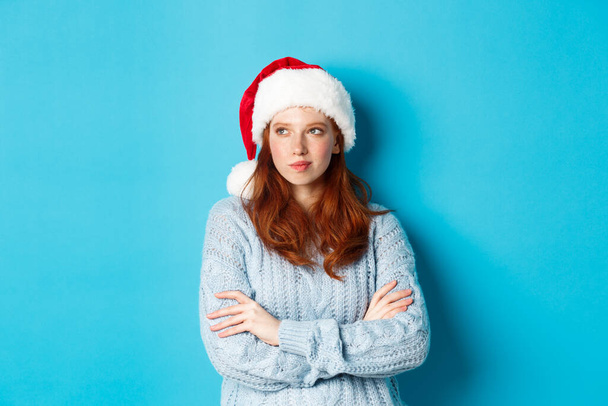 Winterferien und Heiligabend. Nachdenkliche rothaarige Frau mit Weihnachtsmütze und Pullover, die nach links schaut und nachdenkt, Weihnachtspläne schmiedet, vor blauem Hintergrund steht - Foto, Bild