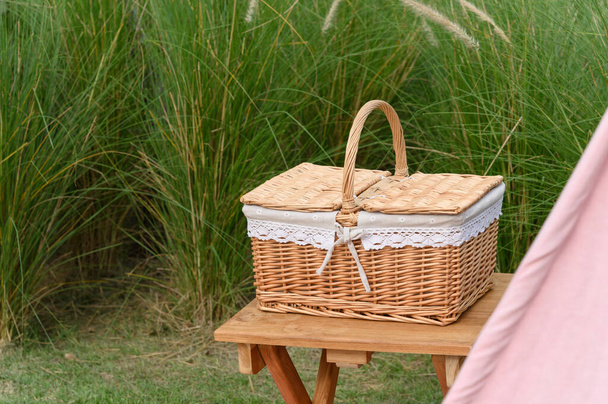 Woven wood picnic basket on small wooden table in park. Summer sunny day and picnic time concept - Photo, Image
