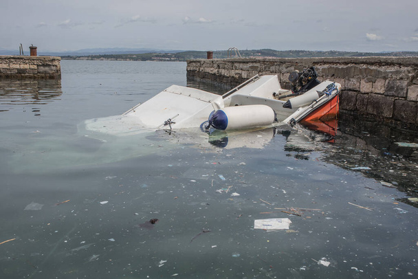 Um pequeno barco de plástico afundou parcialmente sob a água em uma pequena marina perto de um porto de Koper, na Eslovênia. Escombros ou lixo são vistos flutuando na água. - Foto, Imagem