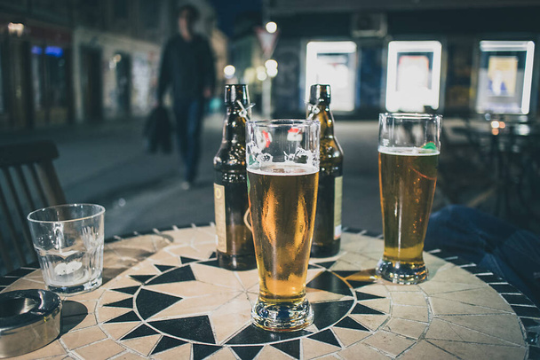 Deux bières lager dans des verres d'un demi-litre sur une table de bar en plein air pendant les heures du soir, avec des bouteilles brunes derrière. Une personne marche vers la table dans une rue la nuit. - Photo, image