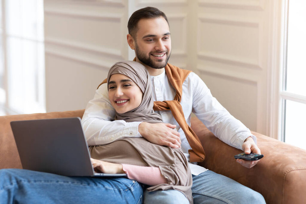 Μουσουλμανικό ζευγάρι χαλαρώνοντας χρησιμοποιώντας Laptop και βλέποντας τηλεόραση στο σπίτι - Φωτογραφία, εικόνα