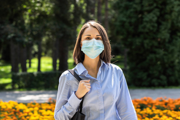 Mujer joven retrato mira a la cámara en un parque de verano con una máscara protectora de la cara médica - Foto, imagen