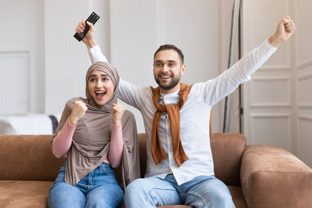 Fröhliches muslimisches Paar schaut fern, gestikuliert und sitzt zu Hause - Foto, Bild