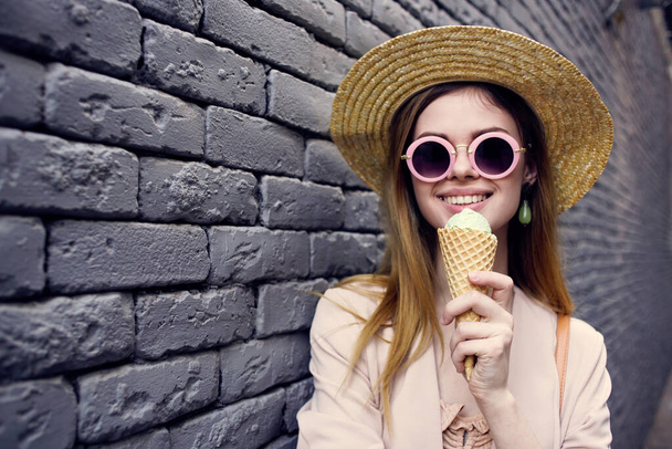 Mulher de óculos de sol e um chapéu com sorvete andando pela parede de tijolos da cidade ao ar livre - Foto, Imagem