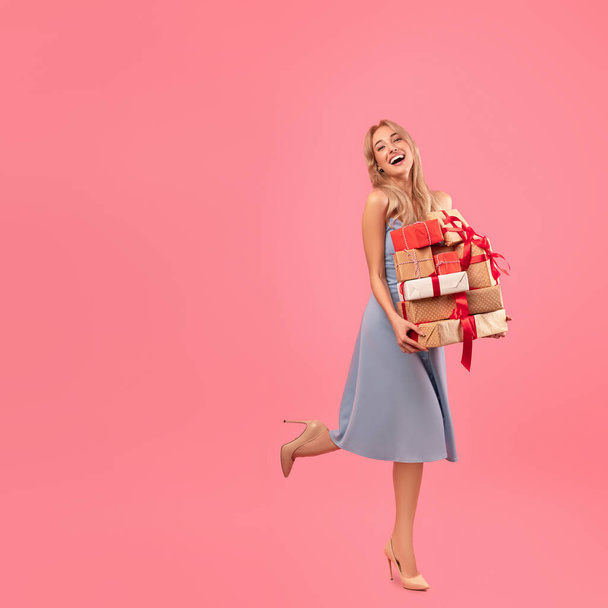 Holiday sale. Attractive millennial lady in pretty dress holding stack of presents on pink background, copy space - Photo, Image