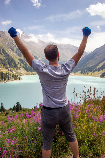 Un hombre levantó las manos al cielo junto a un lago de montaña. Espectacular paisaje en el gran lago Almaty, montañas Tien-Shan - Foto, Imagen