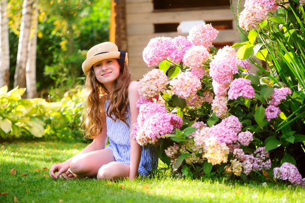 happy kid girl walking in summer garden in straw, posing near pink hydrangea macrofila bush - Photo, image