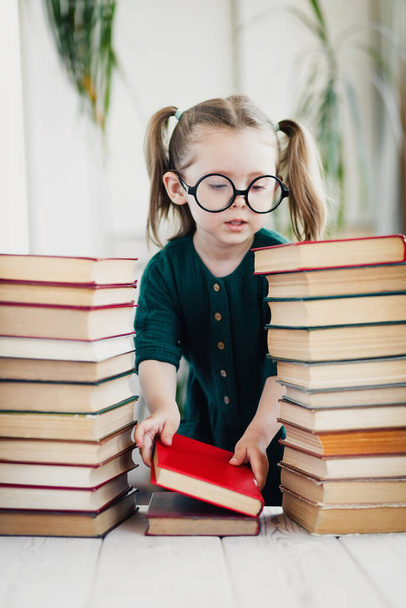 Schattig klein meisje in ronde vorm bril en groene mousseline jurk vouwt kleurrijke boeken in orde - Foto, afbeelding
