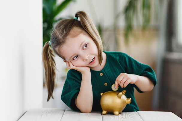 Beautiful cute girl putting money into gold piggy bank at table indoors. Primary financial education - Foto, Bild