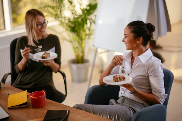 Twee jonge zakenvrouwen die taart eten op kantoor - Foto, afbeelding